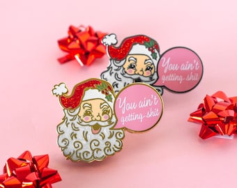 May include: Two Santa Claus enamel pins with gold trim and red glitter hats, each with a pink speech bubble that reads "You ain't getting shit." Red bows are scattered on a pink background. Christmas-themed.