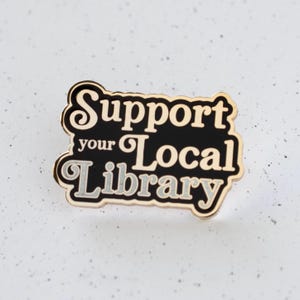 May include: Black and gold enamel pin with the text "Support your Local Library" in white lettering.