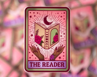 May include: A pink and green tarot card sticker with a crescent moon and a book with a floral design. The text on the sticker reads "The Reader".