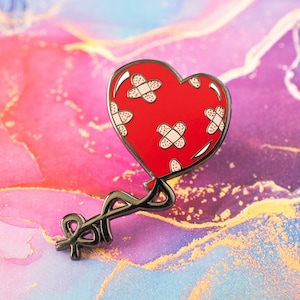 Puede incluir: Un pin de esmalte rojo en forma de corazón con vendas blancas. El pin tiene un contorno negro y un tallo negro con un lazo. El fondo es una colorida mezcla de rosa, morado y dorado.