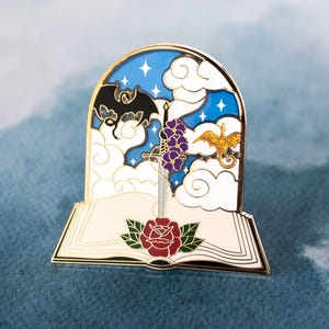 May include: Gold-toned enamel pin featuring a book with a sword piercing through it, a rose, a dragon, and a bat. The book is open and the sword is surrounded by purple flowers. The background is a blue sky with white clouds and stars.