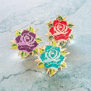 Puede incluir: Tres pines de esmalte con un diseño de rosa. Las rosas son en tonos de rojo, morado y turquesa. Cada rosa está rodeada de hojas verdes.