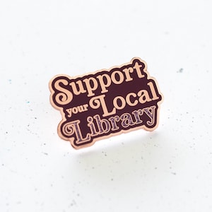 May include: A rose gold enamel pin with the text "Support your Local Library" in a vintage font. The text "Library" is glittery.