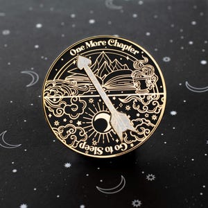 Puede incluir: Un pin de esmalte negro y dorado con una flecha blanca apuntando hacia la derecha. El pin presenta un libro, una montaña, una taza de té, una luna y estrellas. El texto en el pin dice "One More Chapter Go to Sleep".