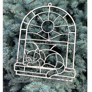 May include: A light-colored wooden cutout ornament featuring an arched window frame design with a circular top section. Inside the frame, a stylized cat is depicted lying down on the window sill. The piece includes a small hole at the top for hanging.