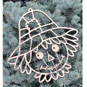 May include: A light-colored wooden cutout of a scarecrow face wearing a wide-brimmed hat with a patch detail. The design features circular eyes, a triangular nose, and a stitched mouth, with a small hole at the top for hanging purposes.