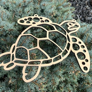 May include: A wooden turtle-shaped ornament with a cutout design. The ornament is light brown and features a detailed shell pattern, head, and flippers. It is designed to be hung and is resting on a bed of blue-green pine needles.