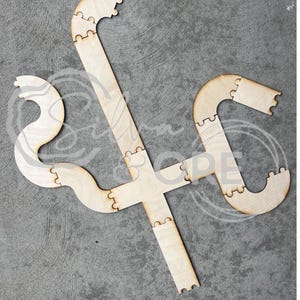 May include: A light-coloured wooden puzzle track composed of multiple interlocking segments. The pieces form a central cross shape with curved, winding extensions branching out from the middle, featuring puzzle-style connectors on the edges.