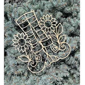 May include: A light-colored wooden cutout featuring a pair of boots with buckled straps, surrounded by two sunflowers and leafy vine accents. The design includes a grid pattern on the boots and small circular holes at the top for hanging purposes.