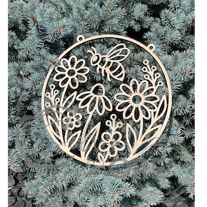May include: A circular light-colored wooden cutout featuring a laser-cut design of a bee surrounded by various stylized flowers and leafy stems. The piece has two small holes at the top for hanging and displays intricate floral patterns.