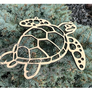 May include: A laser-cut wooden sea turtle wall hanging featuring a geometric shell pattern and circular cutouts on the flippers. The light-colored wood has dark, charred edges from the cutting process, creating a high-contrast outline of the turtle.