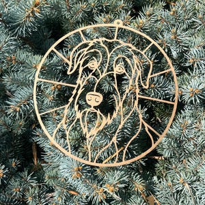 Op de afbeelding: Een cirkelvormig ornament van lichtbruin hout met een lasergesneden omtrek van een hond met een ruige vacht en een baard. Het ontwerp is omsloten door een cirkelvormig frame met stralende lijnen die van de hond naar de buitenrand lopen.