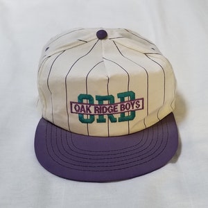 May include: A vintage baseball cap with a purple brim and a cream-colored crown featuring vertical pinstripes. The cap has the embroidered text "ORD OAK RIDGE BOYS" in teal and purple.