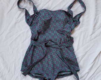 May include: A one-piece swimsuit with a geometric pattern in teal, pink, and white. The swimsuit has a square neckline, shoulder straps, and a tie at the waist. The fabric is patterned with small squares.