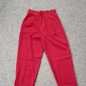 May include: Bright red high-waisted pants with pleats and a button closure. The pants are made of a smooth fabric and have a classic, tailored design. The pants are laid flat on a neutral-colored surface.