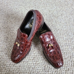 May include: A pair of burgundy leather loafers with a crocodile skin texture. The shoes have a gold tassel detail and the brand name "Netleton" is visible on the inner sole. The shoes are displayed on a neutral-toned woven rug.