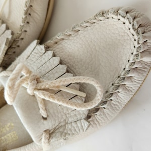 Puede incluir: Un par de mocasines de cuero de color blanco roto con detalles de flecos y un ribete trenzado alrededor del borde. Los mocasines tienen un cord&oacute;n de cuero de color crema.