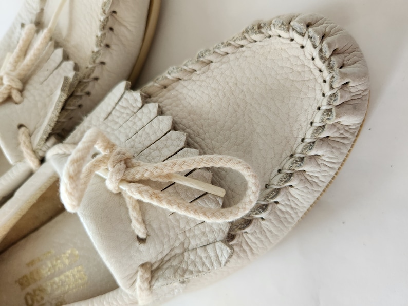 Puede incluir: Un par de mocasines de cuero de color blanco roto con detalles de flecos y un ribete trenzado alrededor del borde. Los mocasines tienen un cord&oacute;n de cuero de color crema.
