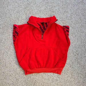 May include: A bright red, sleeveless sweatshirt with a high collar and a half-zip closure. The inside of the collar and the sleeve edges feature a black and brown zebra stripe pattern. The sweatshirt has a front pocket and a ribbed hem.