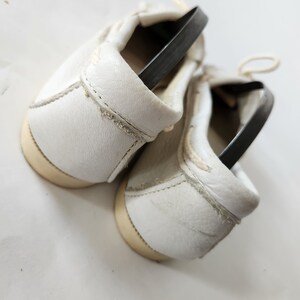 Puede incluir: Un par de zapatos de cuero blanco con correas el&aacute;sticas negras. Los zapatos est&aacute;n atados juntos y los cordones son visibles.