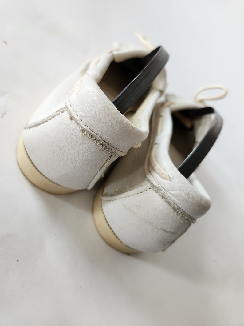 Puede incluir: Un par de zapatos de cuero blanco con correas el&aacute;sticas negras. Los zapatos est&aacute;n atados juntos y los cordones son visibles.