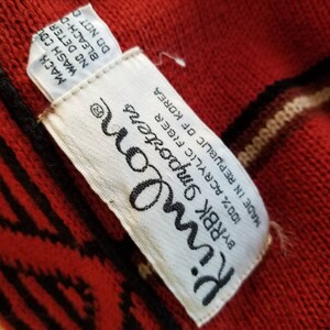May include: A white tag with black text that reads "Kimlone RBK Importer 100% Acrylic Fiber Made in Republic of Korea" attached to a red sweater with black and white stripes.