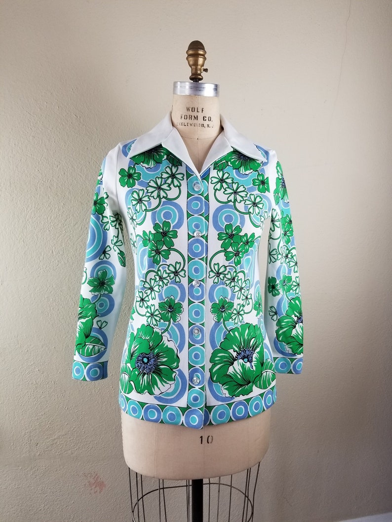 May include: A vintage button-down shirt with a white collar and cuffs. The shirt features a vibrant floral pattern in shades of green and blue on a white background. The long-sleeved shirt is displayed on a mannequin.