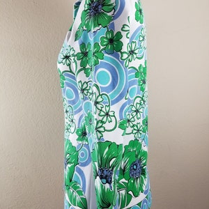 May include: A white blouse with a vibrant floral pattern in shades of green, blue, and white. The design features large stylized flowers and circular motifs. The blouse has a collared neckline and long sleeves, showcasing a retro, vintage style.