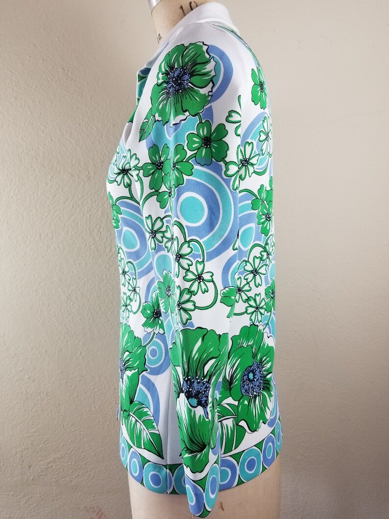 May include: A white blouse with a vibrant floral pattern in shades of green, blue, and white. The design features large stylized flowers and circular motifs. The blouse has a collared neckline and long sleeves, showcasing a retro, vintage style.