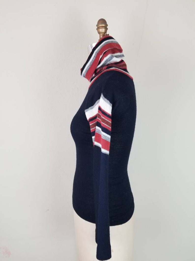 May include: A navy blue long-sleeved turtleneck sweater with red, white, and gray striped detailing on the sleeves and collar.