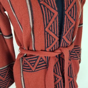 May include: A rust-colored knitted cardigan with a black and white geometric pattern. The cardigan has a tie belt and long sleeves.