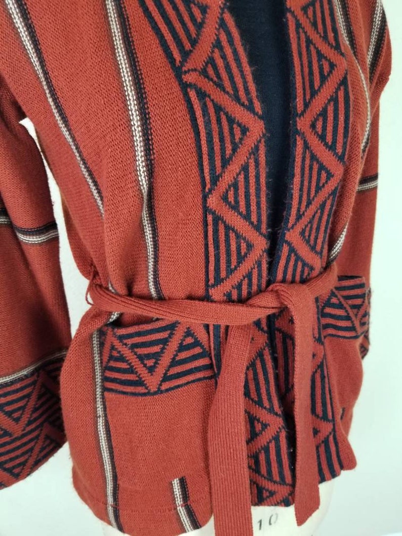 May include: A rust-colored knitted cardigan with a black and white geometric pattern. The cardigan has a tie belt and long sleeves.