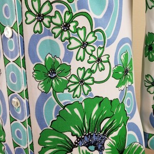 May include: A white garment with a vibrant floral pattern in green, light blue, and teal. The design features large stylized flowers and swirling shapes. The garment has a button-up front with white buttons.