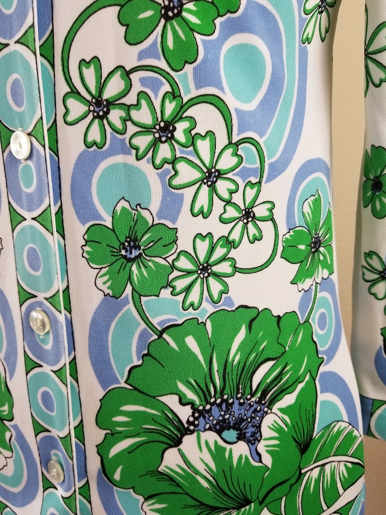 May include: A white garment with a vibrant floral pattern in green, light blue, and teal. The design features large stylized flowers and swirling shapes. The garment has a button-up front with white buttons.