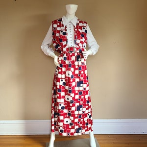 May include: A vintage two-piece outfit on a mannequin. The outfit includes a white collared shirt, a red, white, and blue patterned vest, and a matching floor-length skirt. The skirt and vest feature a geometric square design. The outfit is accessorized with a belt.
