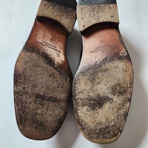 May include: Close-up of the soles of a pair of black leather shoes. The shoes are worn and have a brown leather sole with the text "J.B. Boot New York" and "Made in Brazil" printed on them.