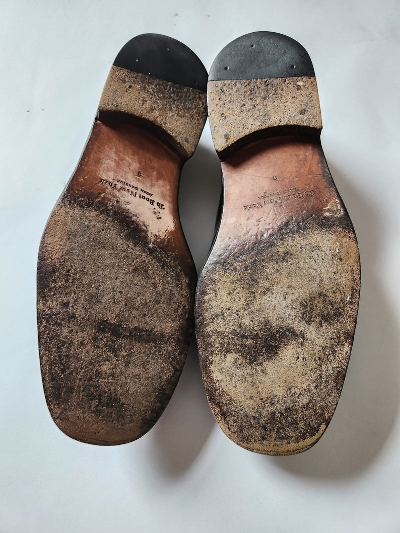 May include: Close-up of the soles of a pair of black leather shoes. The shoes are worn and have a brown leather sole with the text "J.B. Boot New York" and "Made in Brazil" printed on them.