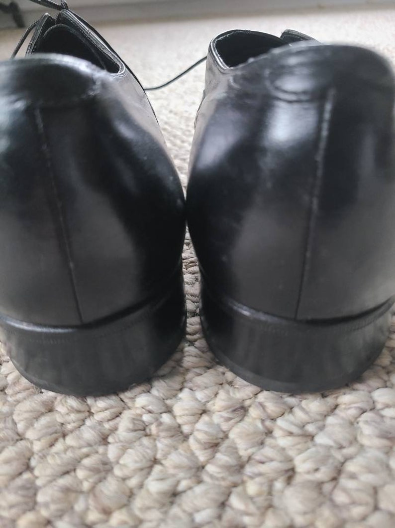 May include: A pair of black leather dress shoes. The shoes have a glossy finish and a classic design, with laces visible at the top. The heels are a solid, dark color.