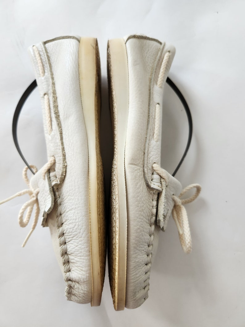 Puede incluir: Un par de zapatos n&aacute;uticos de cuero blanco con cordones blancos. Los zapatos est&aacute;n colocados uno al lado del otro con las suelas mirando hacia el espectador.