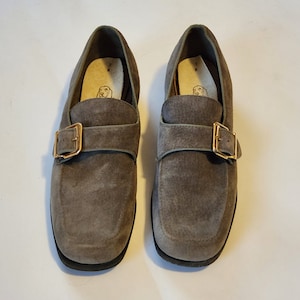 May include: A pair of gray suede buckle loafers with a brown leather strap and buckle. The loafers have a rounded toe and a low heel.