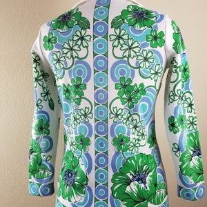 May include: A white long-sleeved top with a vibrant floral pattern in green and blue. The design features large flowers and circular motifs, with a white collar and cuffs. The top is displayed on a mannequin.