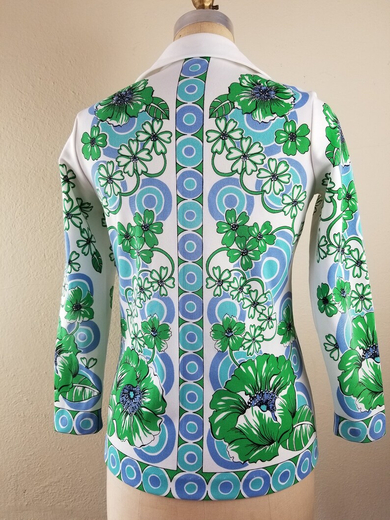 May include: A white long-sleeved top with a vibrant floral pattern in green and blue. The design features large flowers and circular motifs, with a white collar and cuffs. The top is displayed on a mannequin.