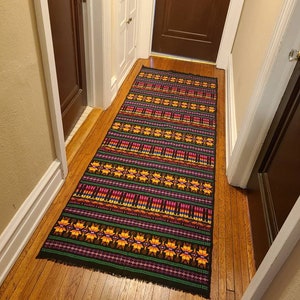 May include: A rectangular, woven runner rug with a black background and a repeating pattern of yellow, pink, green, and purple geometric shapes. The rug is placed on a hardwood floor in a hallway.