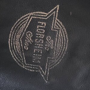 May include: Close-up of a black leather surface with a gold-colored logo. The logo features the text "The Florsheim Shoe" inside a stylized lightning bolt shape, with additional text "Chgo".