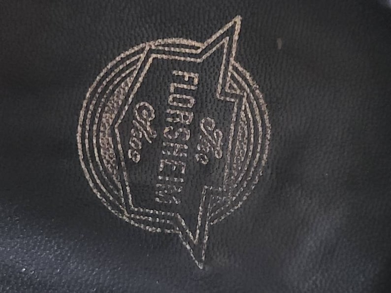 May include: Close-up of a black leather surface with a gold-colored logo. The logo features the text "The Florsheim Shoe" inside a stylized lightning bolt shape, with additional text "Chgo".