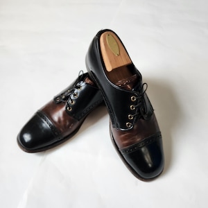 May include: A pair of two-tone leather dress shoes. The shoes are black and brown with a cap toe design and lace-up closure. Wooden shoe trees are inside the shoes. The shoes are on a white background.