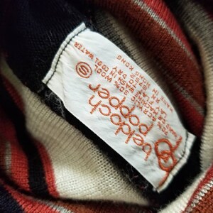 May include: A white tag with black text that reads "2 popper beldoch S 70% &CRYLIC 30% WOOL O MACHINE WASH IN COOL WATER LAY FLAT RN 8752 FLAT TO DRY MADE IN HONG KONG" attached to a striped sweater with red, black, and white stripes.