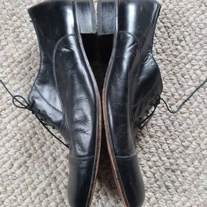 May include: A pair of black leather dress shoes with laces. The shoes have a classic design with a rounded toe and a small heel. The shoes are placed side by side on a textured surface.