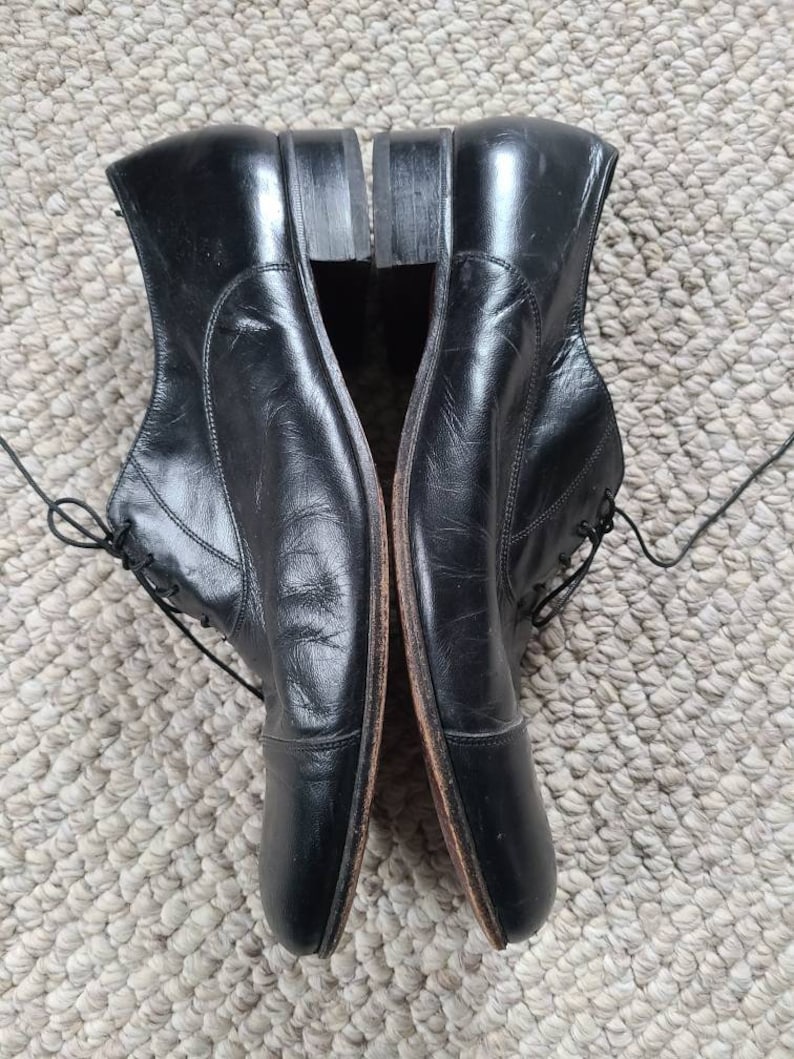 May include: A pair of black leather dress shoes with laces. The shoes have a classic design with a rounded toe and a small heel. The shoes are placed side by side on a textured surface.