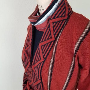 May include: A rust-colored knitted cardigan with a black and white geometric pattern. The cardigan has a shawl collar and a tie belt.